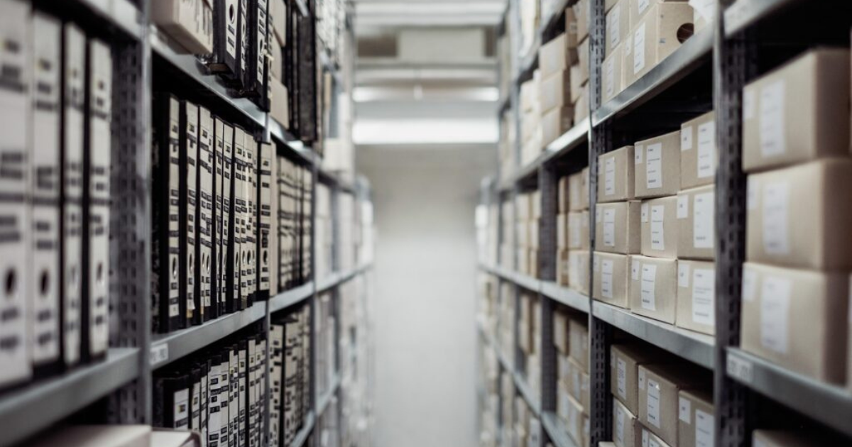 Zdjęcie przjścia między regałami archiwum lub biblioteki z segregatorami i opisanymi kartonami na półkach