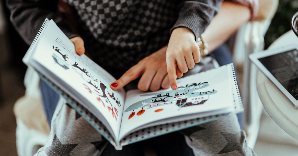 Zdjęcie dziecka siedzącego na kolanach osoby dosrosłej, czytają książkę z obrazkami wskazując ba coś w książce.